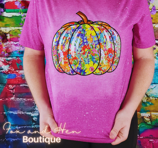 The Design on the t-shirt is a contour of a pumpkin that has a pattern of multicolored flowers. The shirt is a Berry Purple/Pink color with a bleach tie-dye effect. Displayed at a front faced angle. Model is wearing a Large unisex fit.