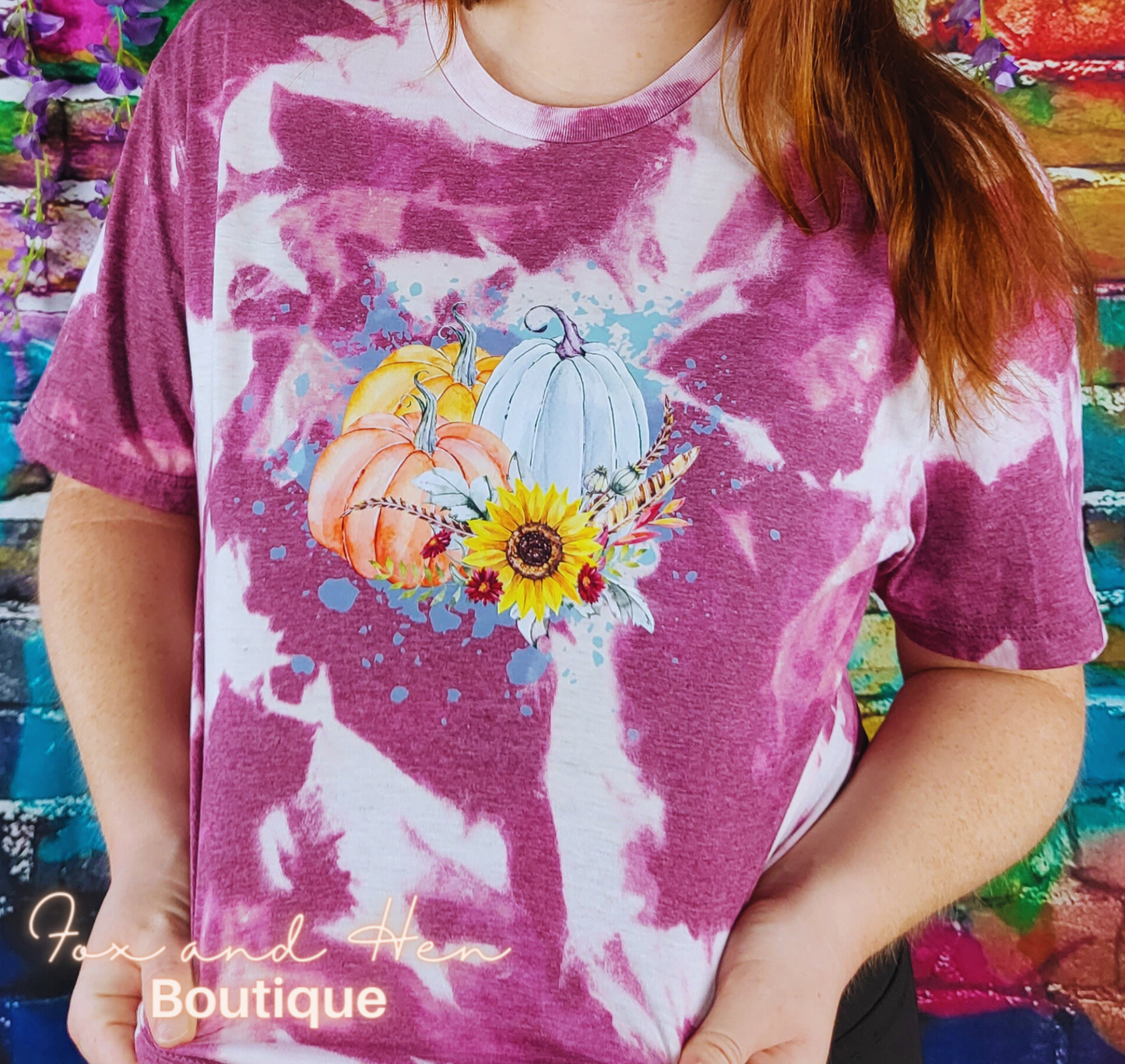 Three multicolored pumpkins with flowers and feathers on top of a paint splattered background on a t-shirt. The shirt is a maroon color with a bleach tie-dye effect. Displayed at an angle. Model is wearing a Large unisex fit.