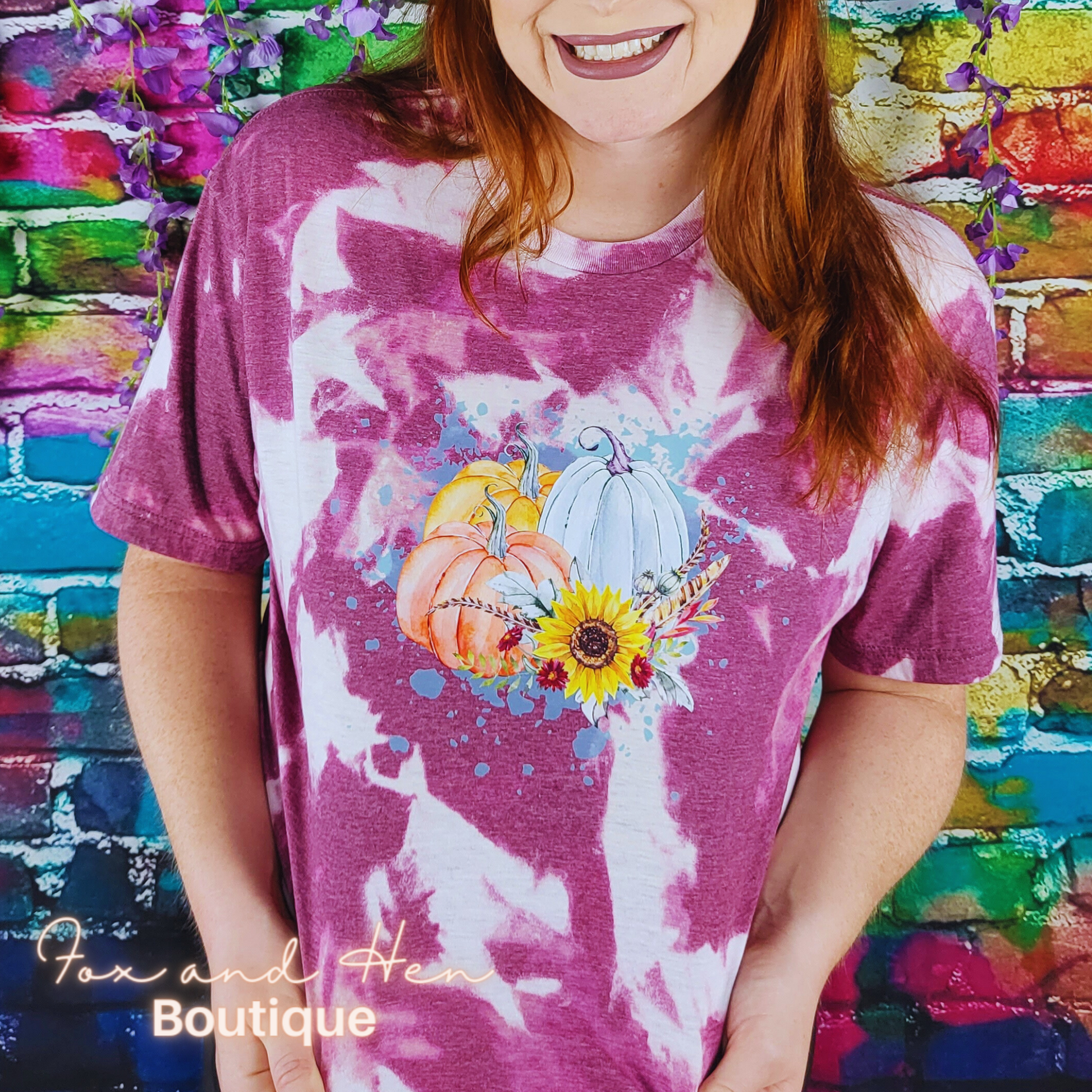 Three multicolored pumpkins with flowers and feathers on top of a paint splattered background on a t-shirt. The shirt is a maroon color with a bleach tie-dye effect. Displayed at a front faced angle. Model is wearing a Large unisex fit.