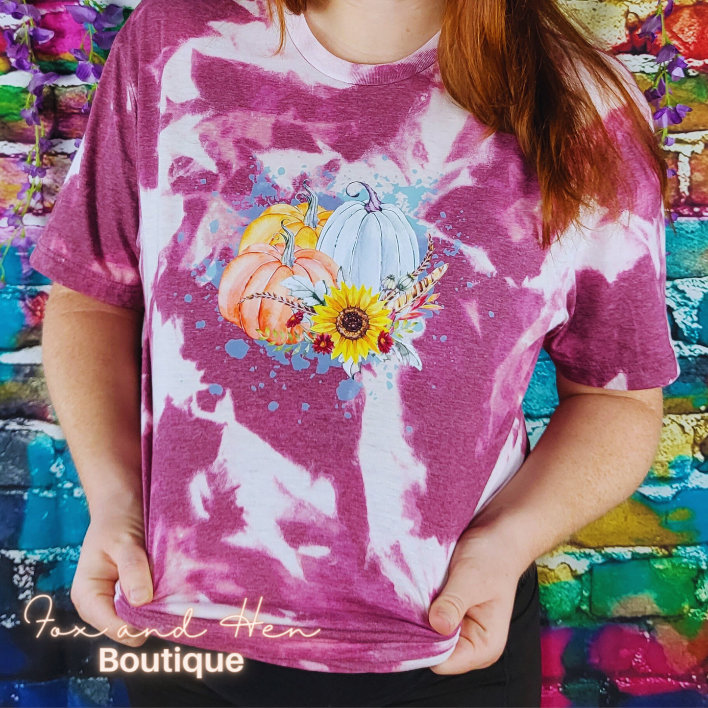 Three multicolored pumpkins with flowers and feathers on top of a paint splattered background on a t-shirt. The shirt is a maroon color with a bleach tie-dye effect. Displayed at a front faced angle. Model is wearing a Large unisex fit.