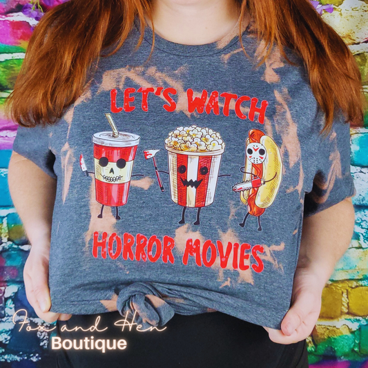 The design on the t-shirt is a cartoon of a soda cup, popcorn bucket, and a hotdog with faces, all carrying weapons, marching to the right. There is text above and below the image in red that reads, "Let's Watch Horror Movies". The shirt is a dark gray color with a bleach tie-dye effect. Displayed at a front faced angle. Model is wearing a Large unisex fit. The model is pregnant and styled the shirt with a knot at the front, showing that it can be worn with a baby bump.